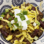 A colorful bowl of chili with grated cheese, sour cream, and green onions